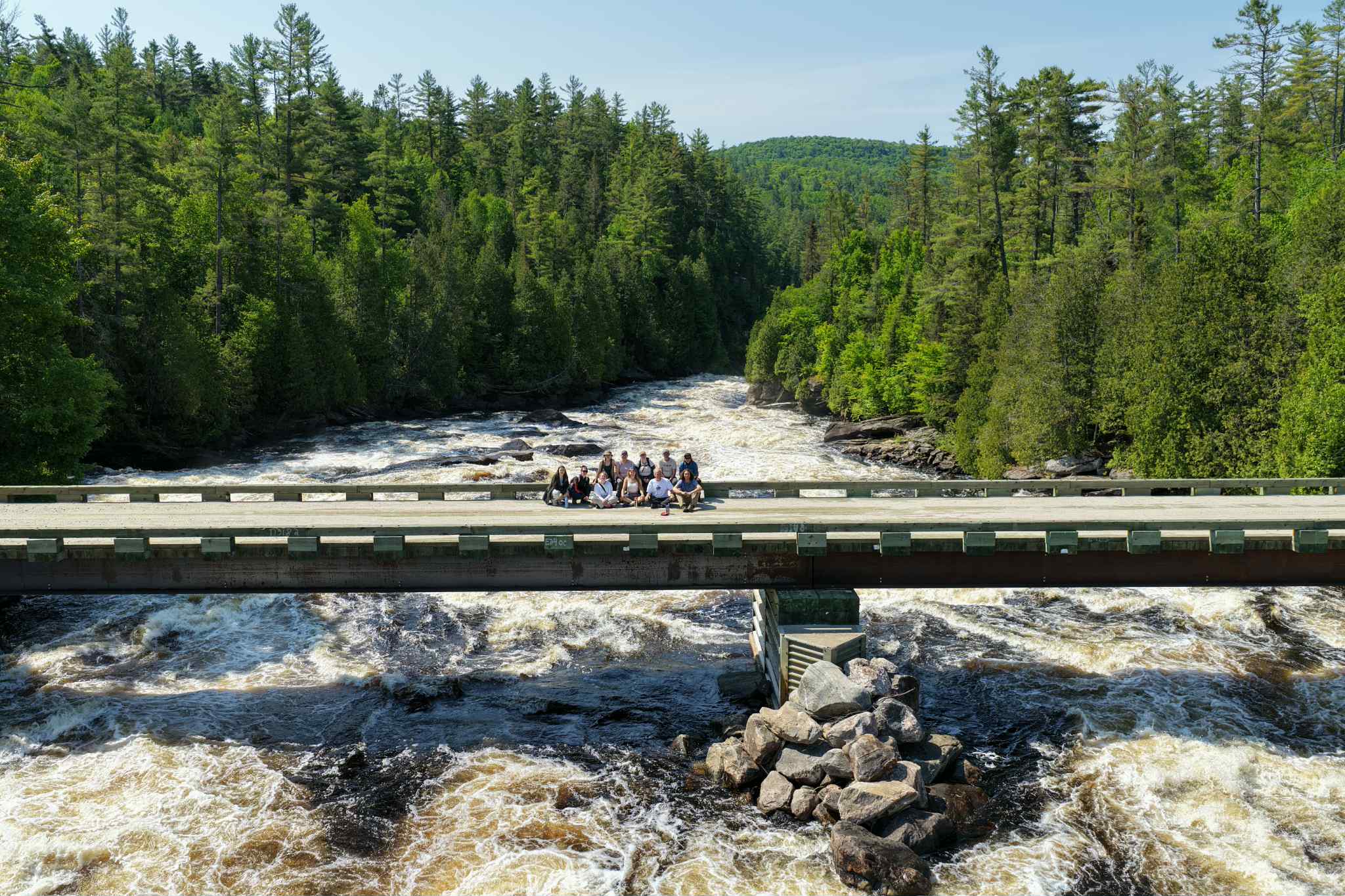 A group of youth and CPAWS-OV staff pose for a photo on a bridge overlooking whitewater rapids surrounded by evergreen forest. They are waving to the camera.